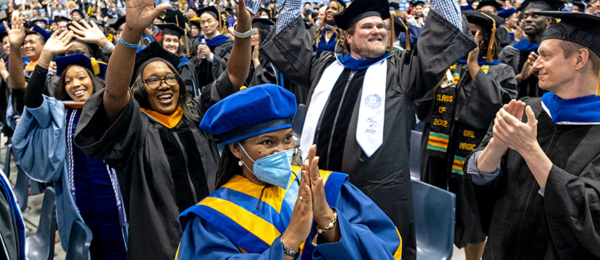 A crowd of doctoral graduates wearing doctoral academic regalia smile and clap as they celebrate their graduation.