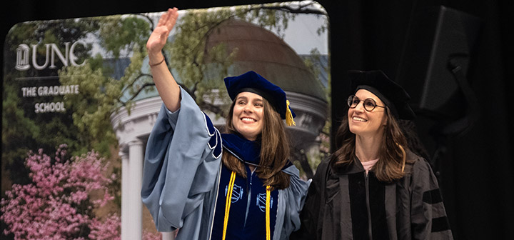 A graduate student in Carolina Blue regalia smiles and waves while standing beside her advisor at the Doctoral Hooding Ceremony