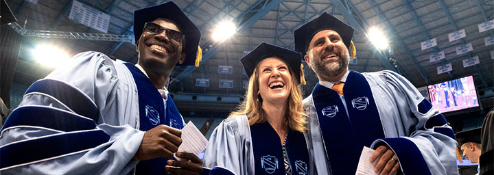 Three graduate students in Carolina Blue doctoral regalia smile broadly at the Doctoral Hooding Ceremony