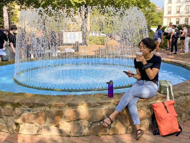 A woman sits on a stone wall surrounding a blue fountain. She is sipping a coffee and looking at her phone.